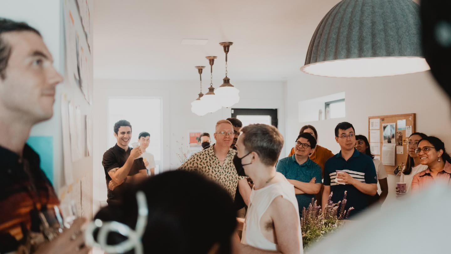 A group of people in a bright room smiling and listening to a speaker. There are hanging lights above and plants visible in the foreground, creating a warm, inviting atmosphere.