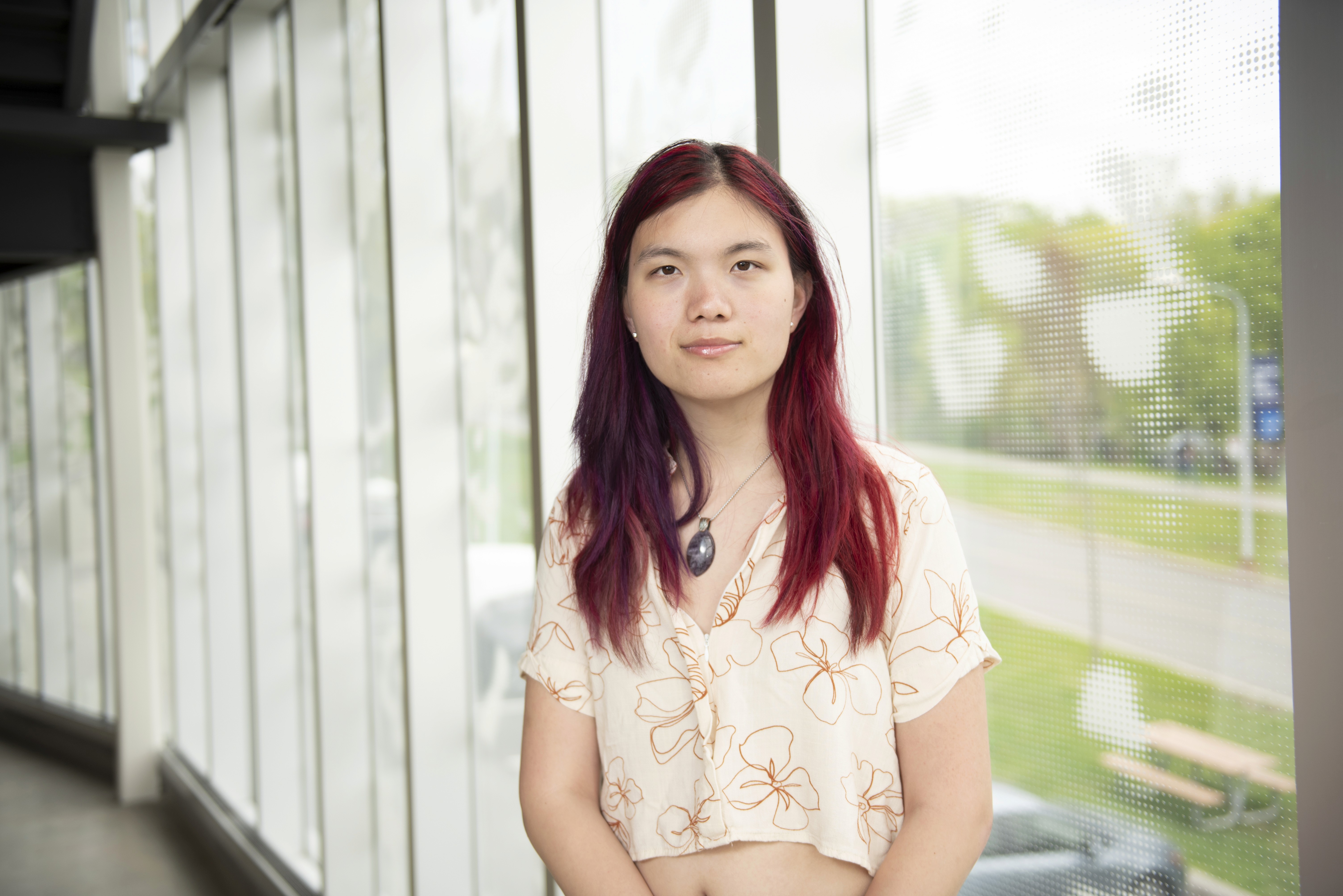 A young person with long hair, standing indoors near large windows, looking directly at the camera.