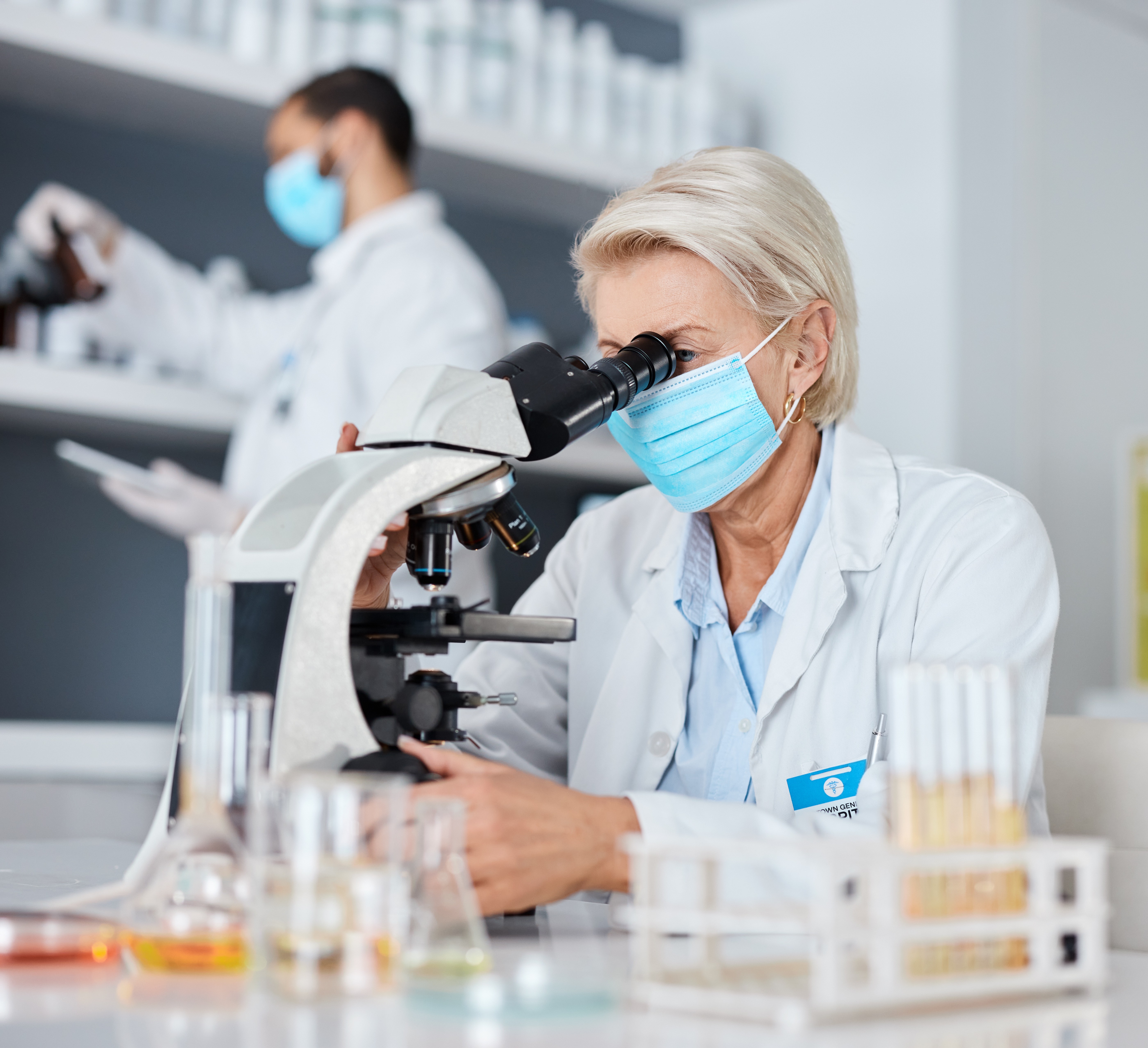Experienced scientist in a lab coat and face mask using a microscope in a research laboratory, indicative of the meticulous talent and dedication in the scientific community.