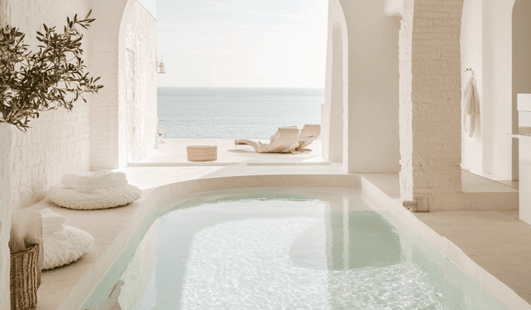 Serene indoor pool with ocean view, white arches, and sun loungers outside.
