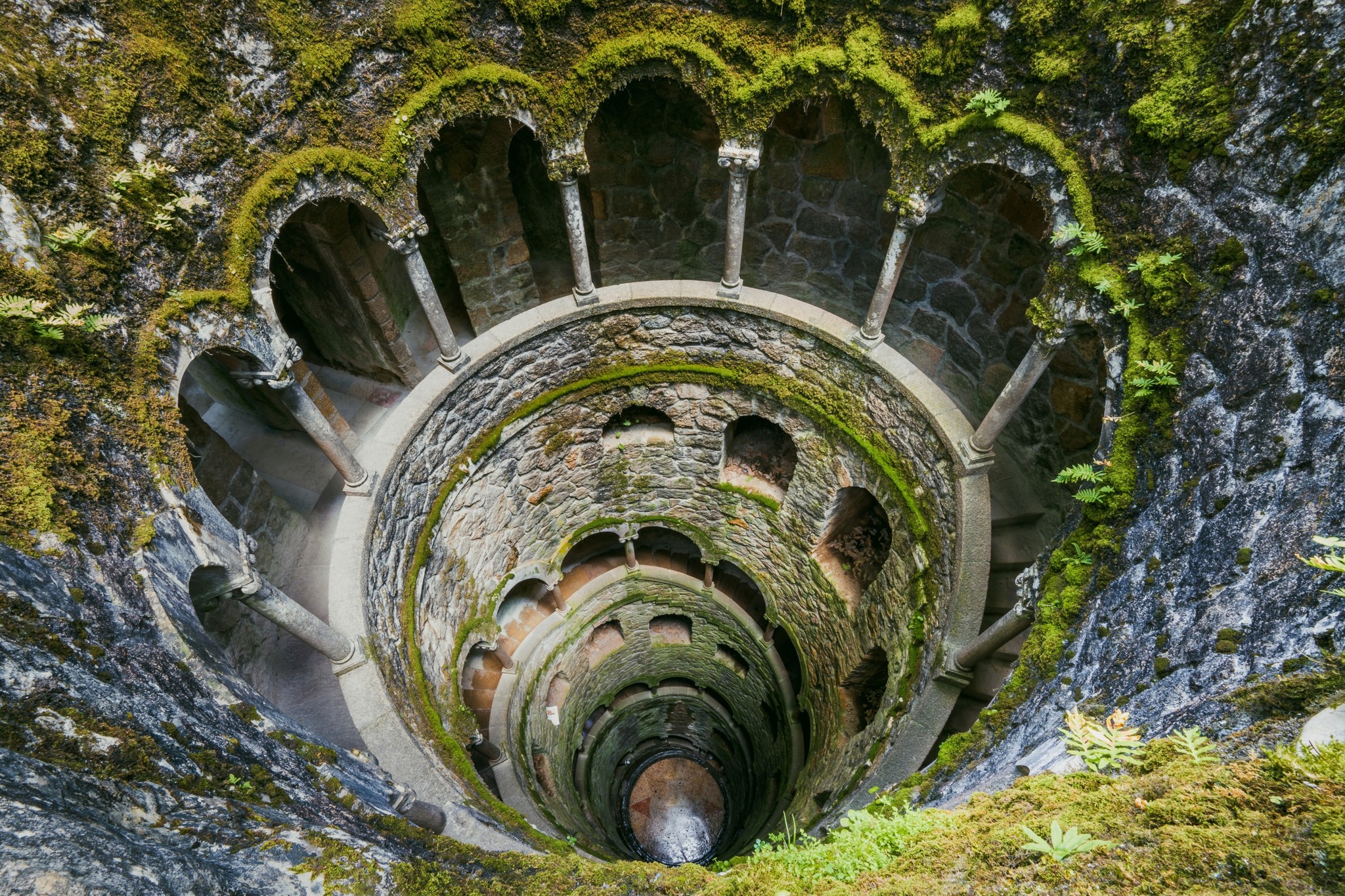 Moss-covered spiral stone staircase tunneling into the ground at Quinta de Regaleira, Sintra, Portugal
