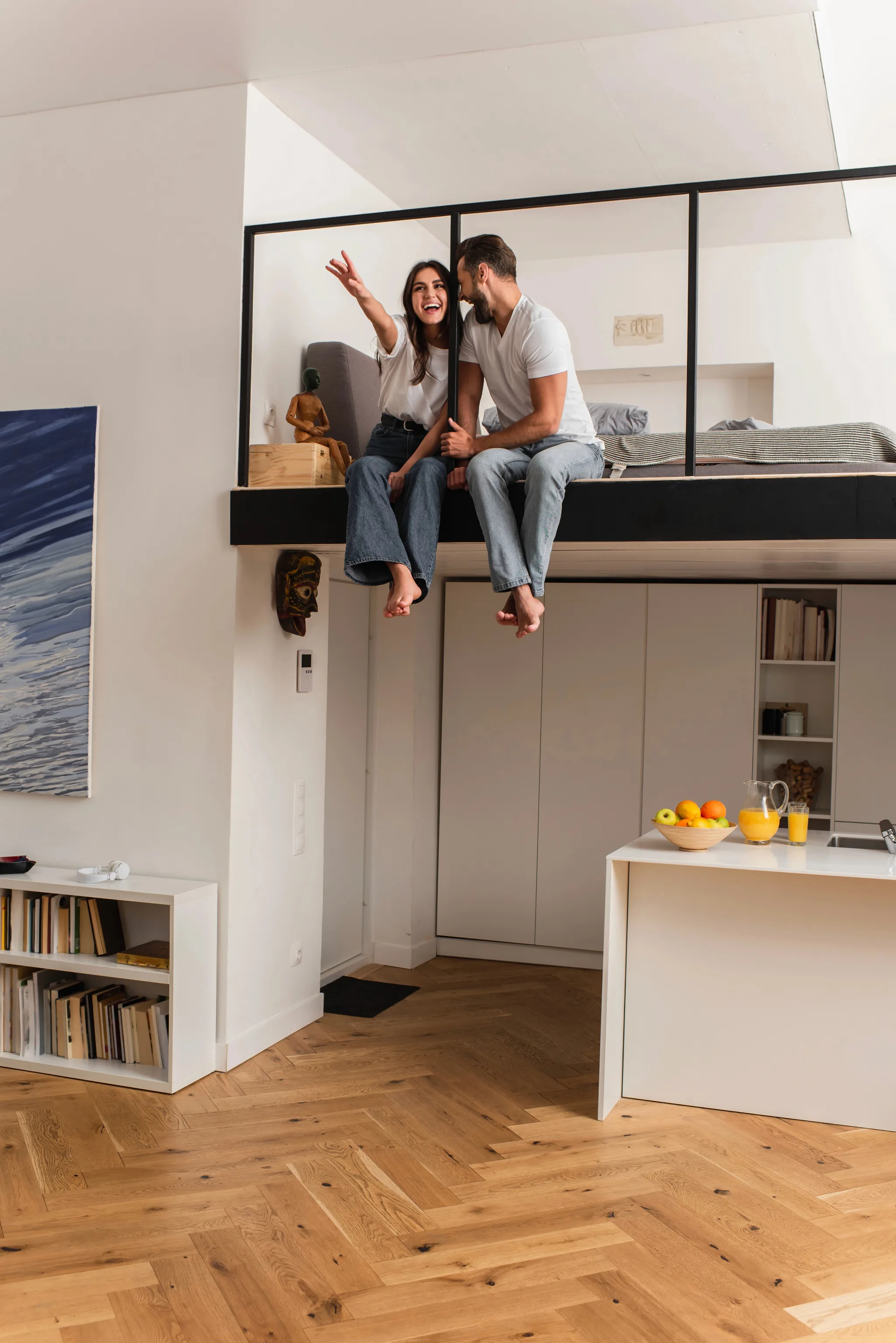 A couple sits together on a lofted platform with black framing in a modern studio apartment, with the woman gesturing while speaking to the man, overlooking a kitchen area below with herringbone wood flooring.