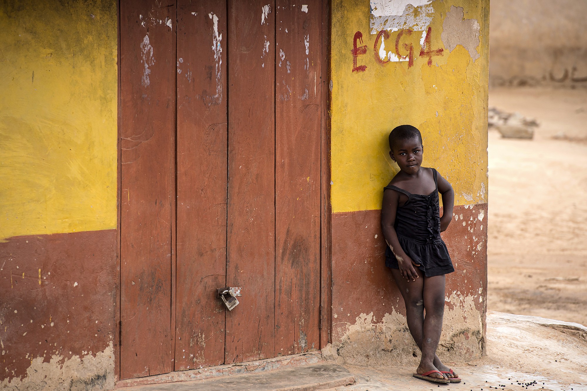 Jeune fille appuyée contre un mur jaune et rouge dans un village, photographiée par Frédéric Bourcier lors d’un reportage documentaire.