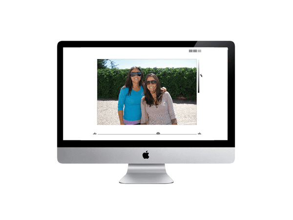 An Apple iMac computer displays a photo on its screen. The photo shows two women standing outdoors in front of a hedge. The woman on the left wears a blue shirt and sunglasses; the woman on the right wears a light-colored top and sunglasses. The photo is centered on the computer screen, and the computer’s silver stand and Apple logo are visible at the bottom.