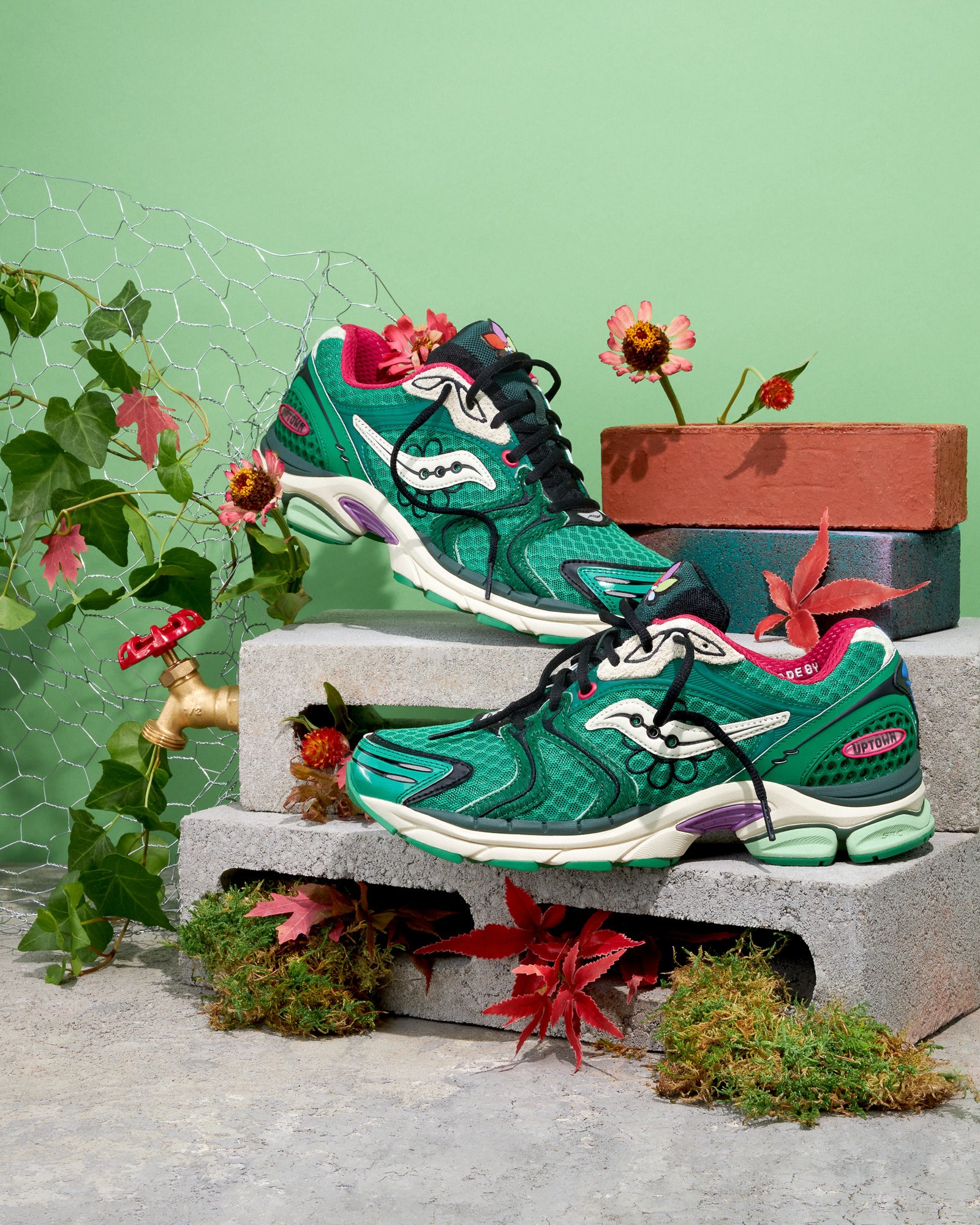 Green Jae Tips x Saucony sneaker on cinder blocks with flowers, moss, and a brass outdoor faucet prop.