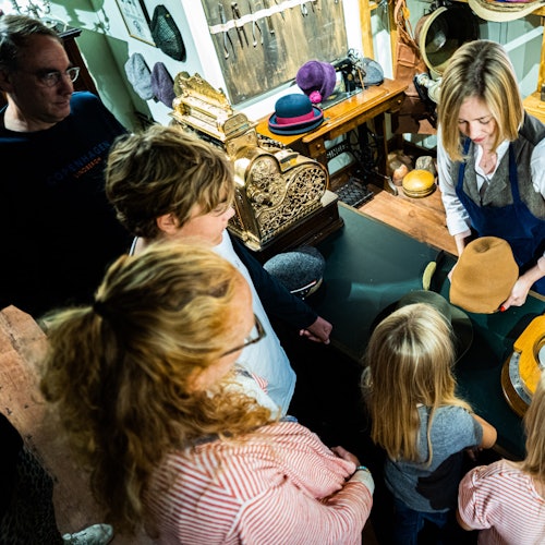 Sekelompok orang, termasuk anak-anak, berkumpul di sekitar seorang wanita yang mendemonstrasikan pembuatan topi di toko vintage.