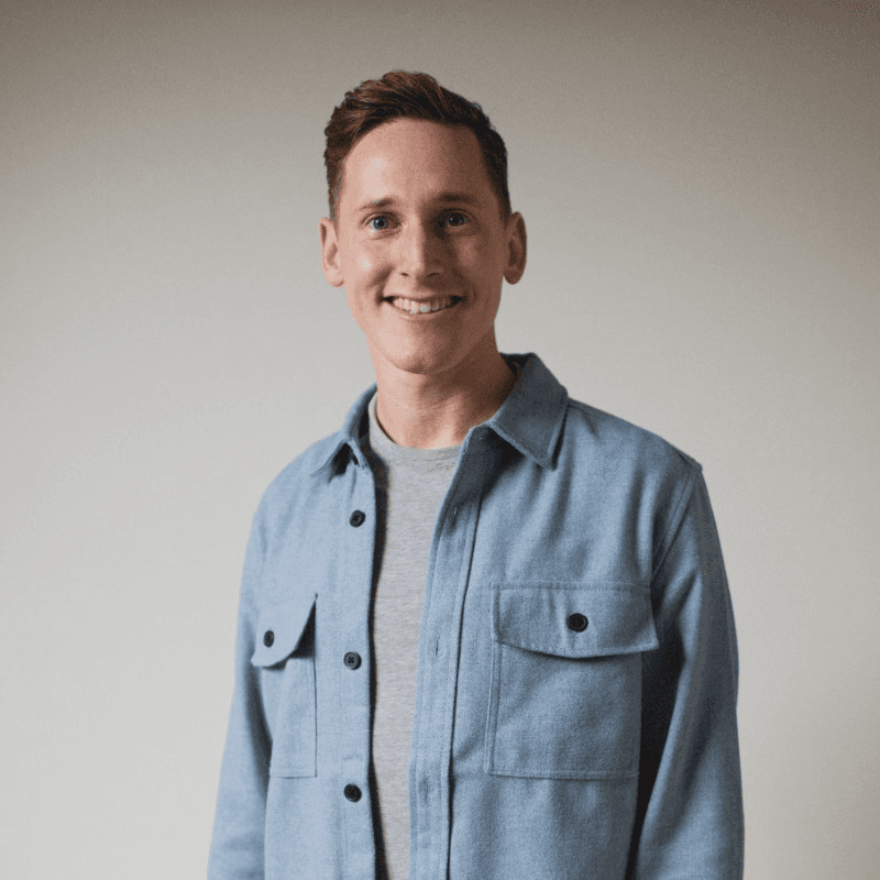 A smiling person wearing a denim jacket stands against a neutral background, showing a friendly demeanor.