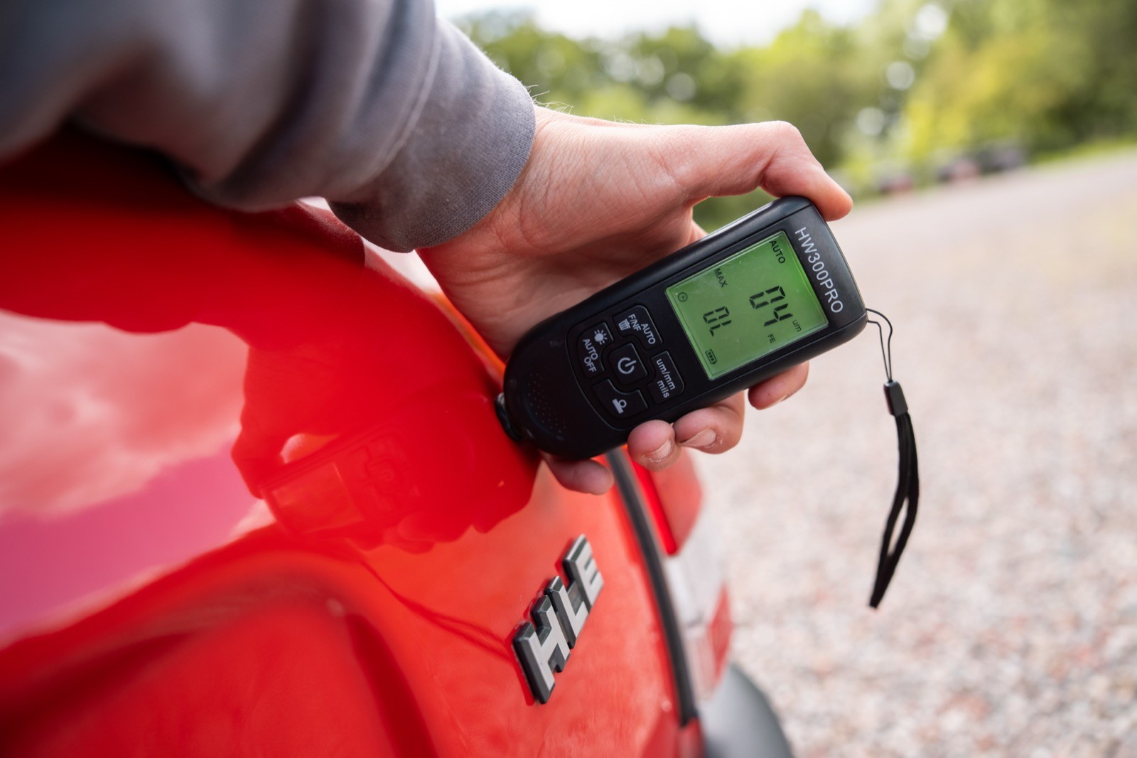 A person holds a device displaying a digital readout, positioned near the taillight of a red vehicle outdoors.