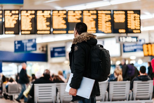 A traveler with a backpack stands in an airport, looking at flight information displays in a busy terminal.