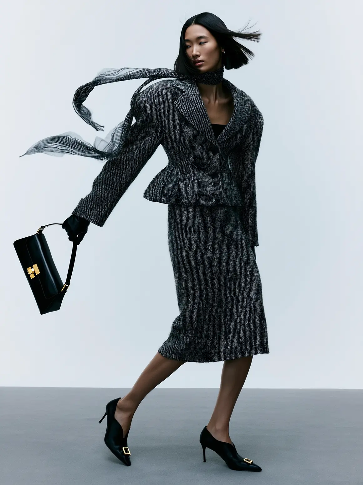 A model elegantly strides in a high-fashion ensemble featuring a tailored charcoal gray suit and matching skirt, accessorized with a black handbag and heeled shoes, embodying the sophisticated style of a high-end fashion brand.
