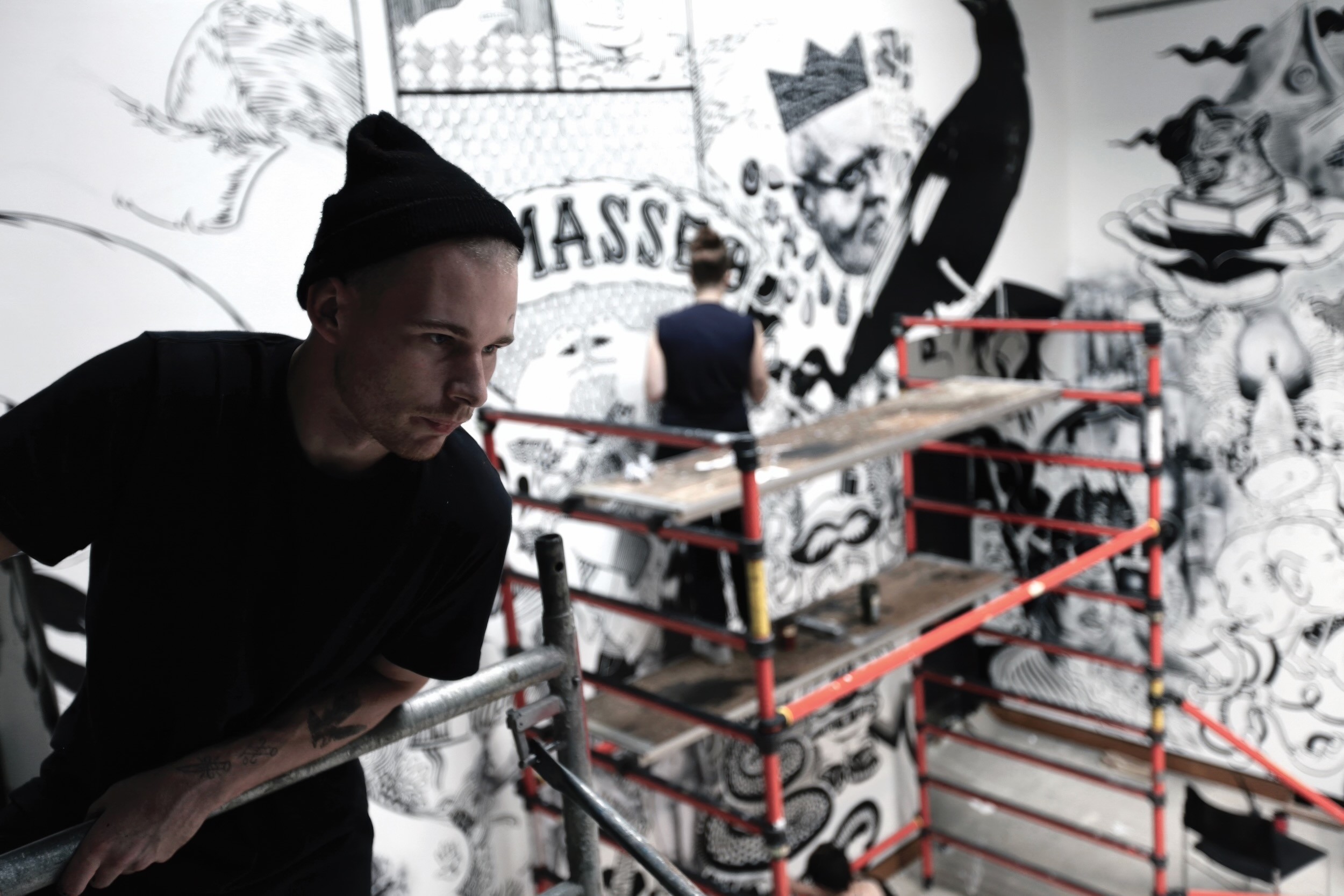 A man in a black knit cap leans on a scaffold rail as another figure draws high on a gallery wall crowded with black-and-white figures and lettering.
