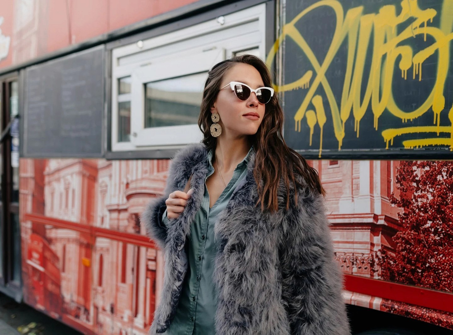 A stylish individual in sunglasses and a faux fur coat poses confidently in front of a colorful street food truck adorned with urban graffiti, showcasing urban fashion.