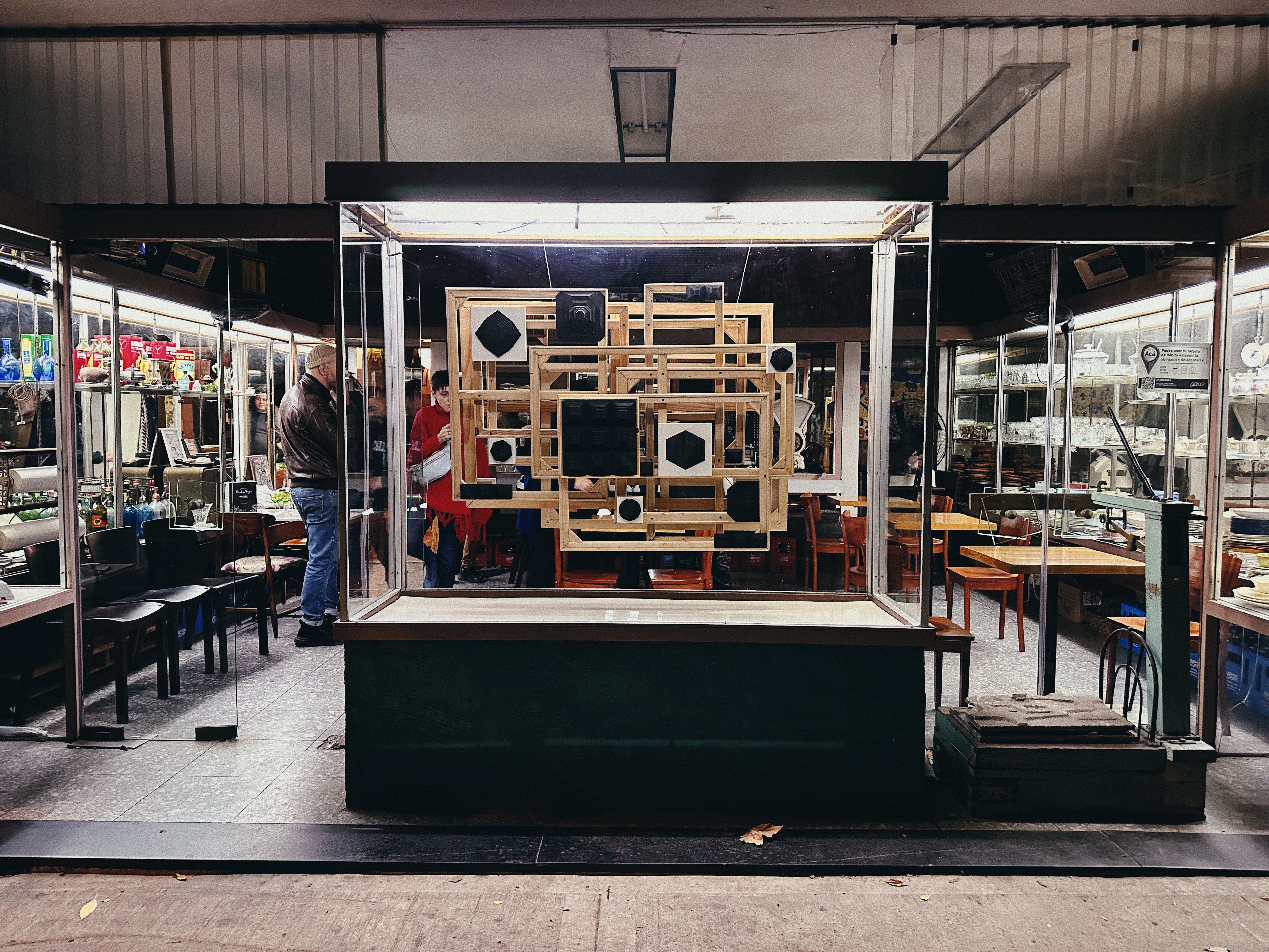 Presenta la vitrina del local, visto desde la calle, con los marcos de madera que son parte de una instalación artística. La imagen capta el reflejo de personas dentro y fuera del local.