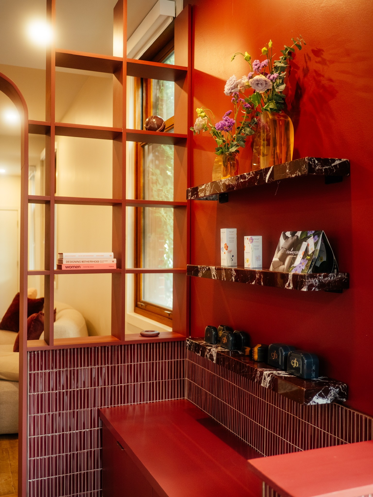 Red marble shelves on a red wall displaying fresh flowers and skincare products, with a glass-paneled screen and burgundy-tiled counter.