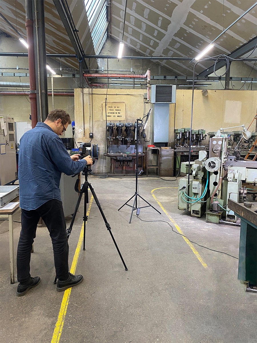Dans un atelier industriel, un homme vêtu d'une chemise bleue et d'un pantalon noir utilise un appareil photo monté sur un trépied pour prendre des clichés. Il fait face à une zone de production où se trouvent des machines-outils lourdes, notamment des perceuses à colonne alignées contre le mur du fond. Un éclairage sur pied est positionné entre le photographe et les machines, et le sol en béton est marqué par des lignes jaunes délimitant les zones de travail.