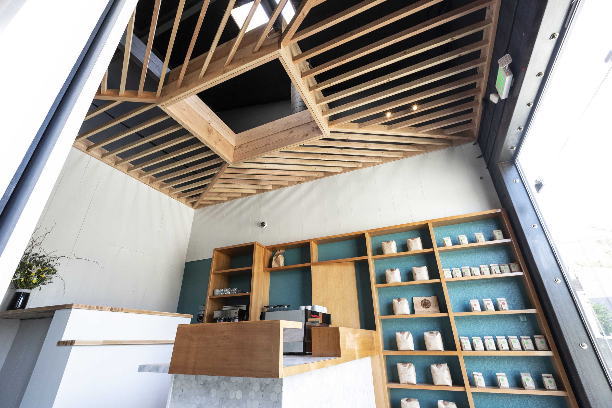 Low-angle view of geometric timber ceiling slats above a coffee counter and teal-backed display shelves.