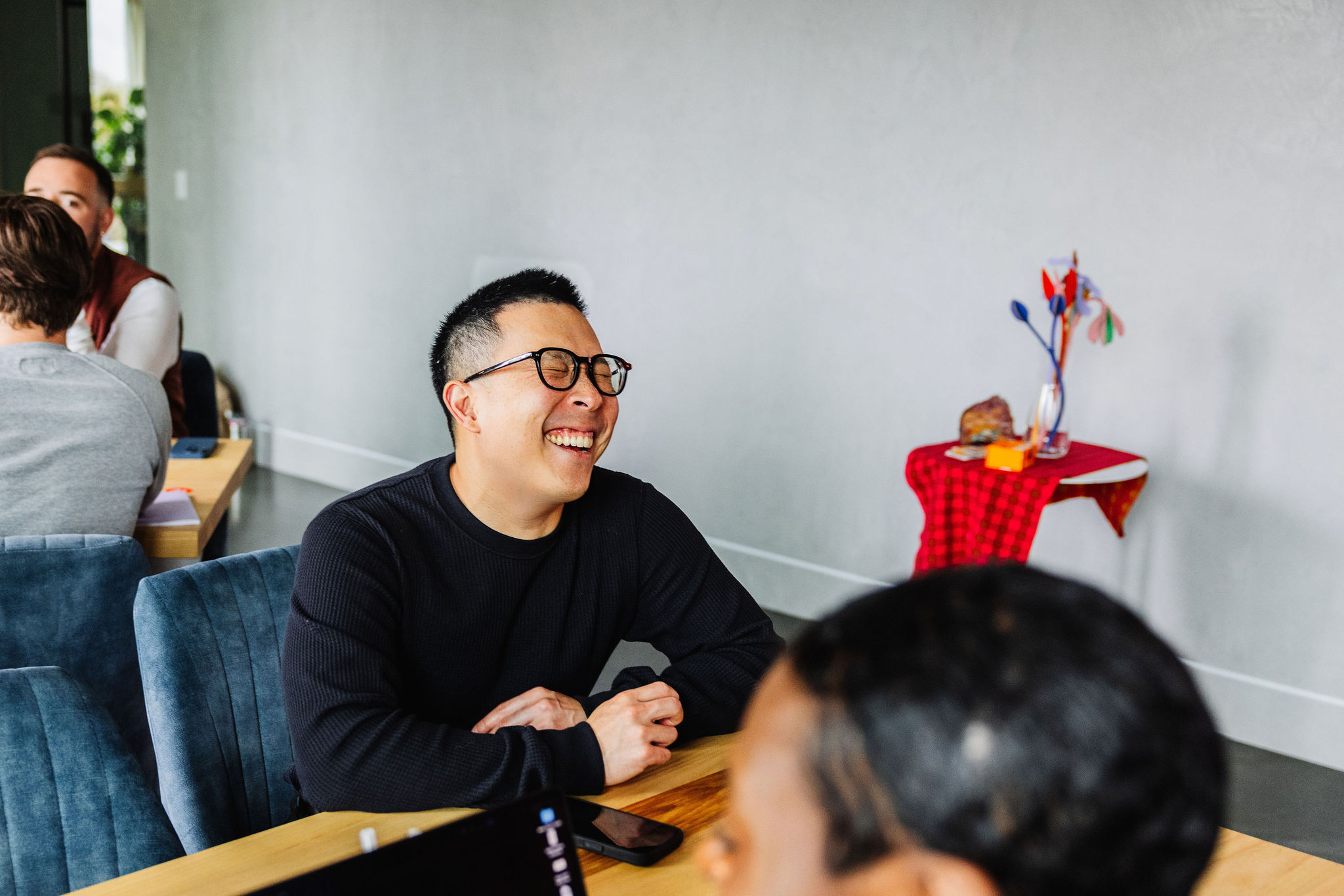 Man laughing while seated at a table during a casual meeting, with others blurred in the background.