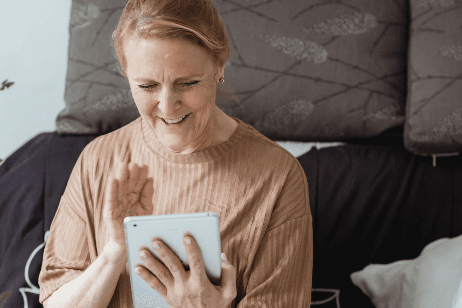 An older woman laughing and waving at a tablet propped in her hands during a video call.