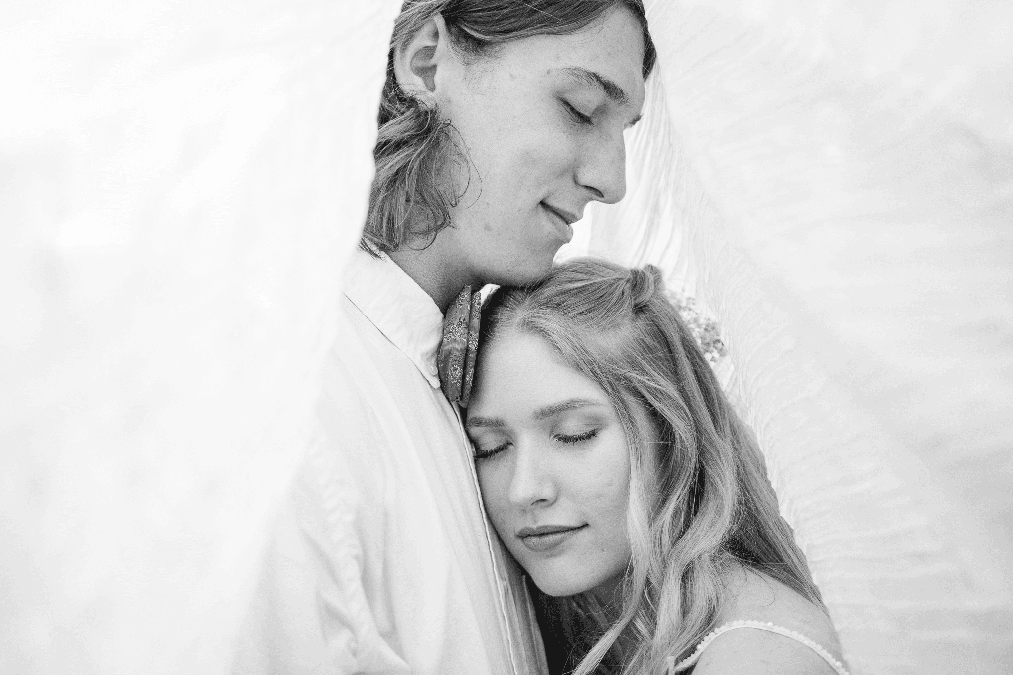  a wedding couple under a veil with their eyes closed with the woman gently placing her head on his chest