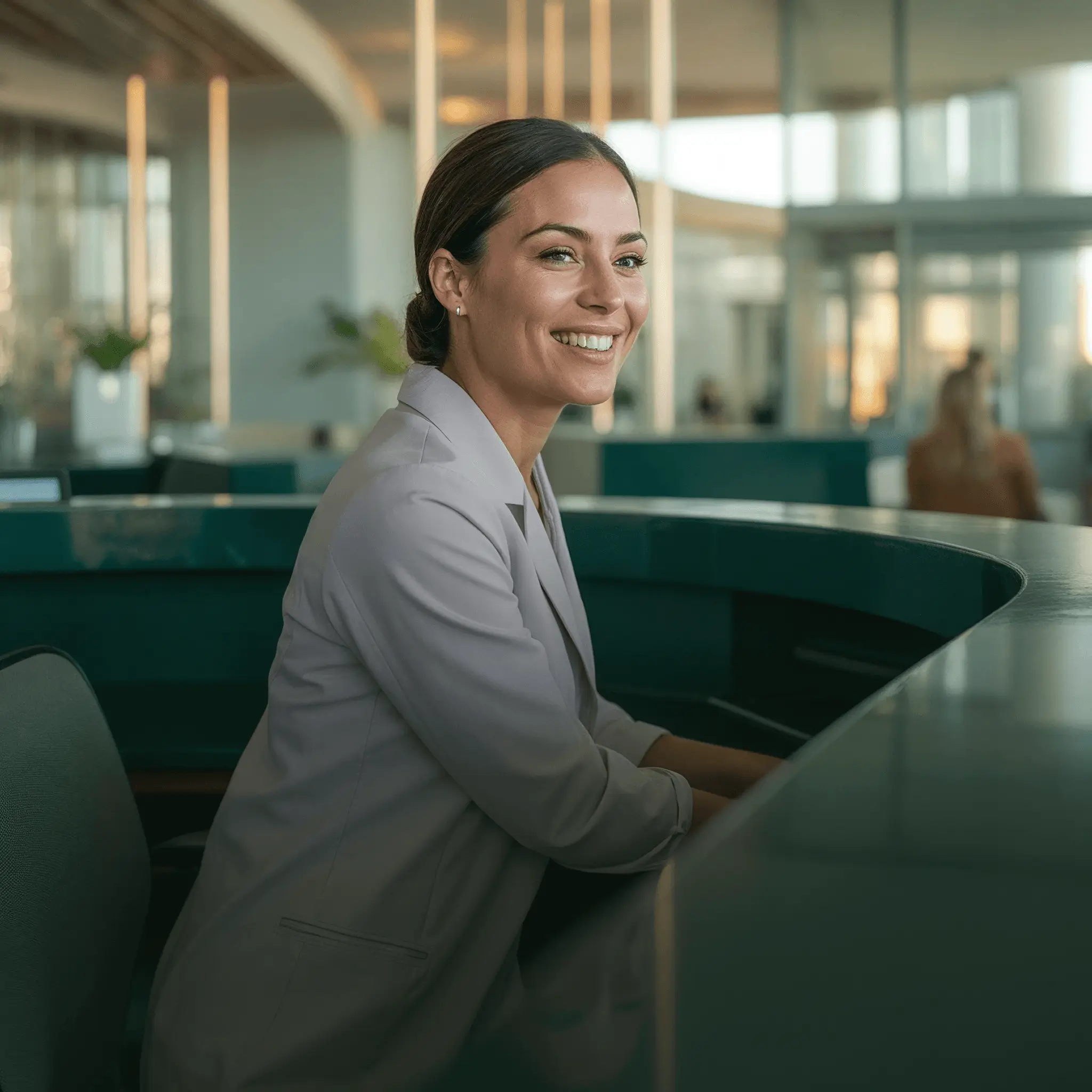 A smiling woman in a light blazer leaning on a curved reception desk in a modern office, with soft natural light and blurred background.