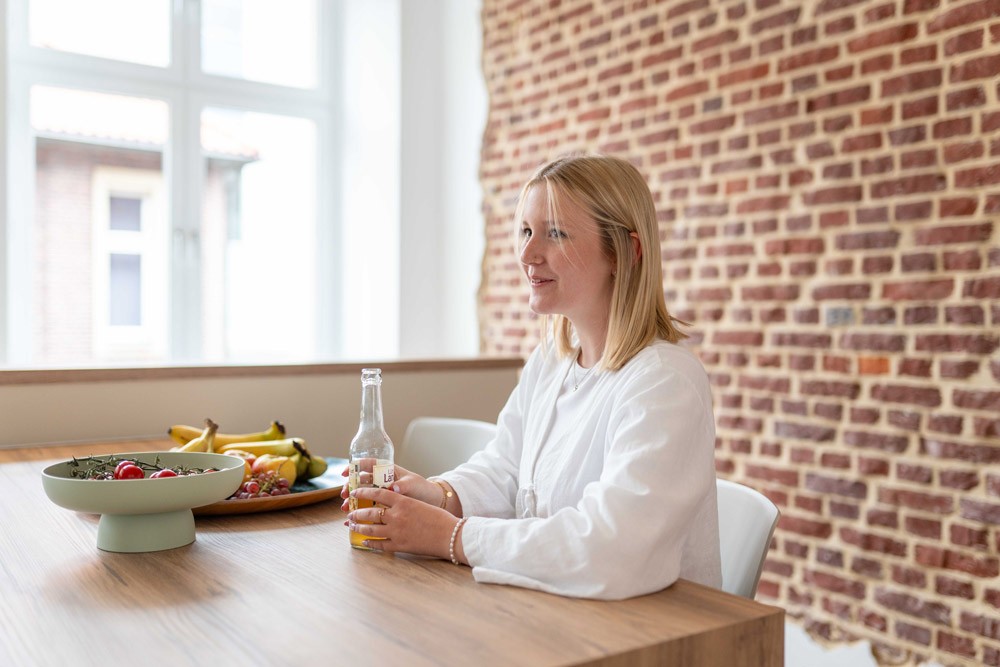 Mitarbeiterin sitzt mit einem Getränk an einem Fensterplatz und denkt nach.