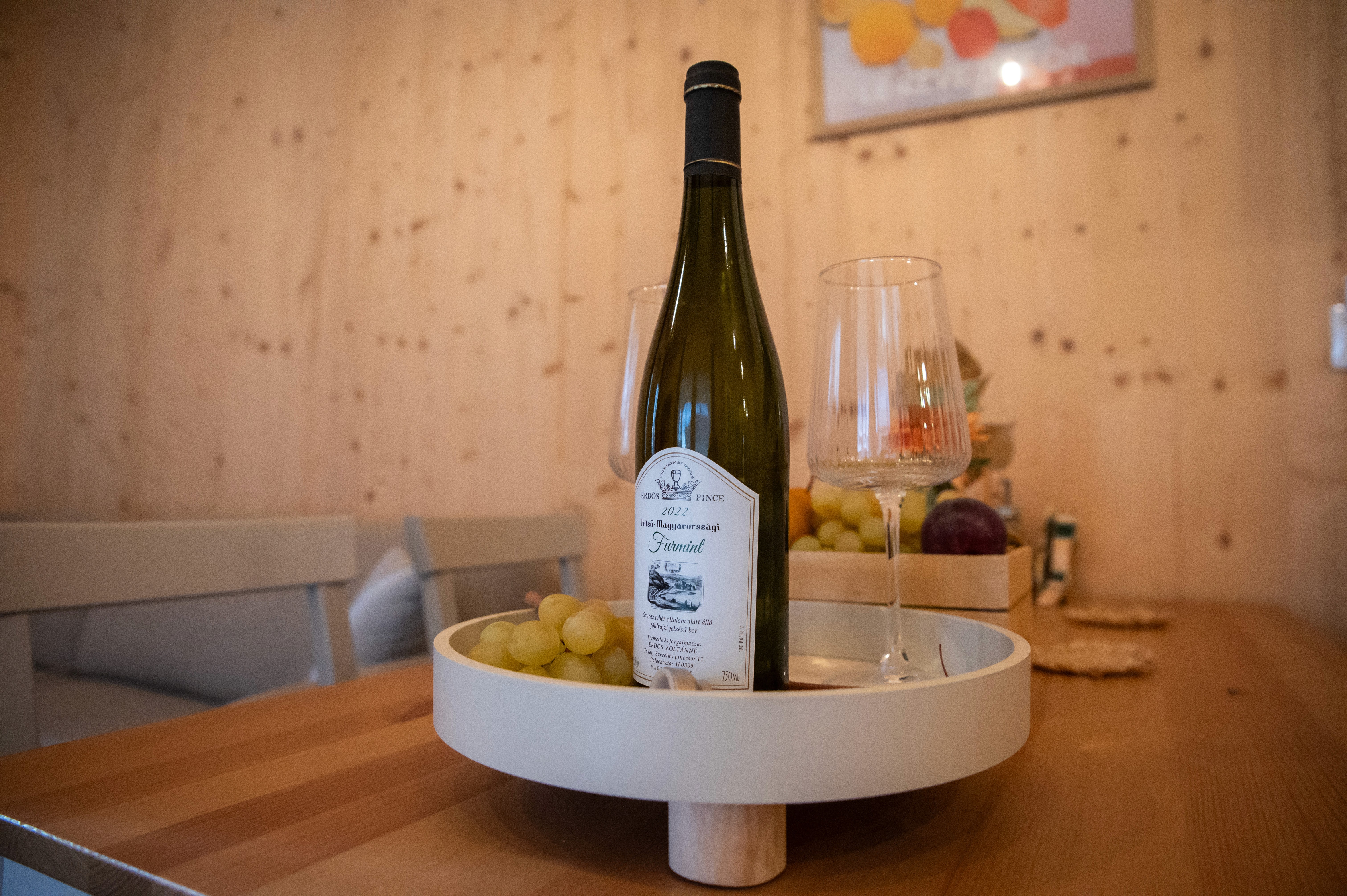 Egy palack fehérbor elegánsan van elrendezve két borospohárral és egy tányér zöld szőlővel egy faasztalon, világos fa fal előtt, meghitt étkezési hangulatot teremtve.