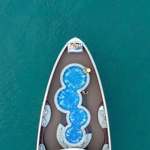Luftfoto af stævnen på en yacht med en unik, opdelt swimmingpool og to personer, der slapper af ved den i det klare blå vand.