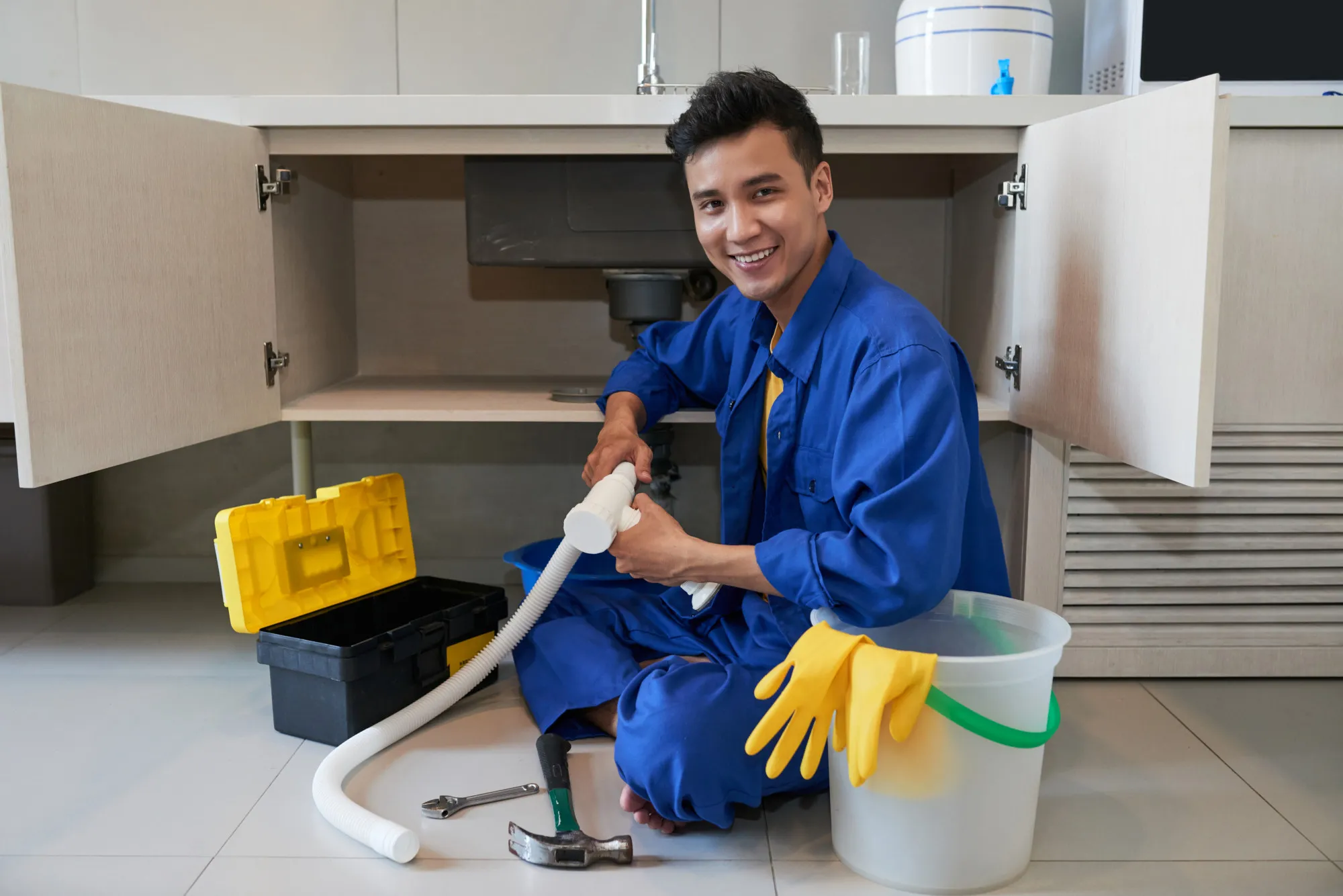 A professional plumber in blue overalls smiling while repairing a kitchen sink drainage system with a toolbox and cleaning supplies nearby.