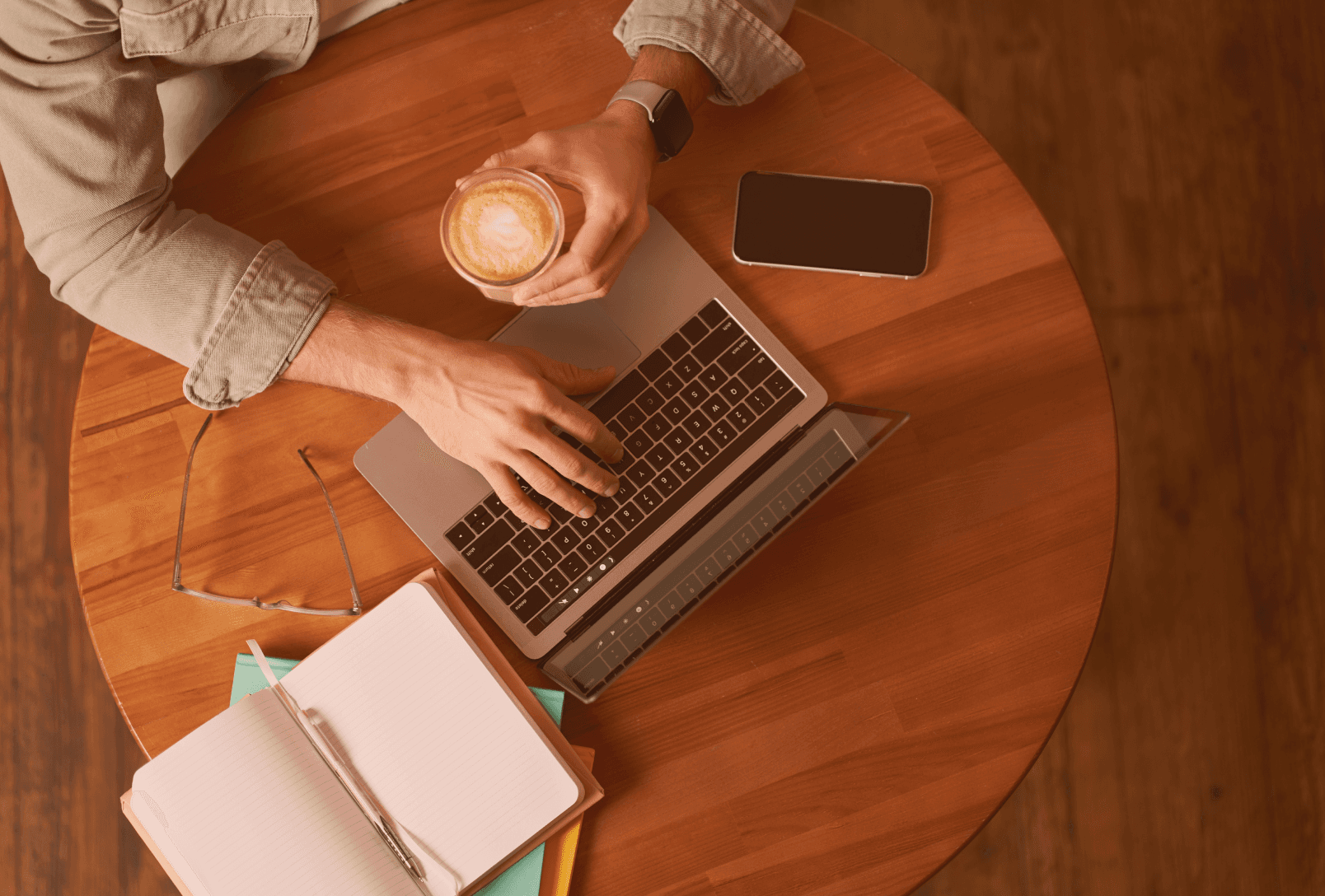 An overhead view of a marketing professional working on a laptop at a wooden cafe table with a latte, notebook, and smartphone, symbolizing a focused workspace and efficient digital workflow.