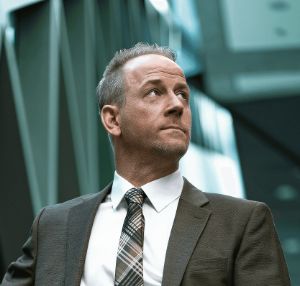 A confident, middle-aged man in a suit and tie stands against a backdrop of modern, geometric architecture, conveying professionalism and leadership.