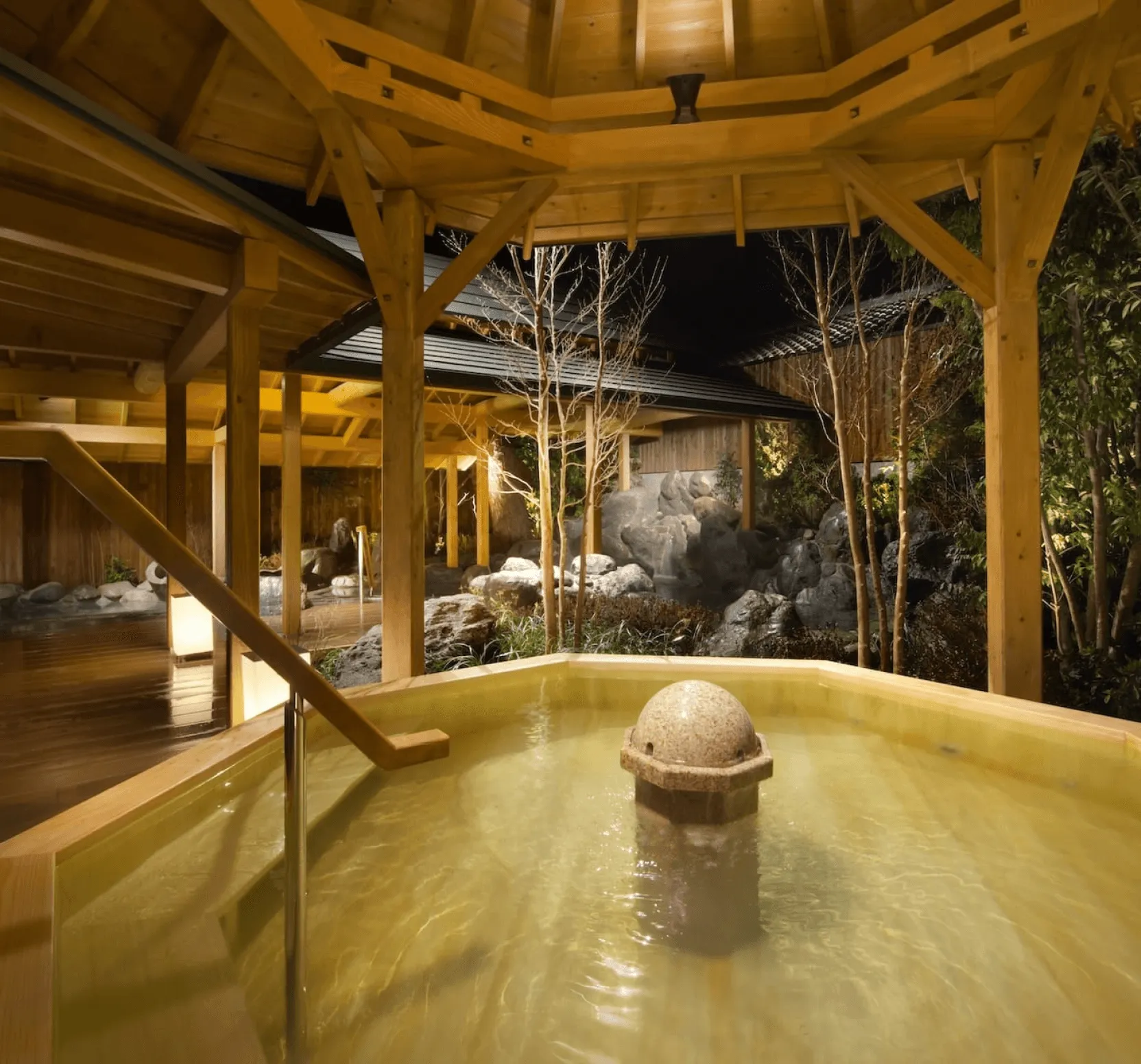 An elegant, multi-sided wooden onsen bath sits under a timber pavilion at night. The entire scene is warmly illuminated, highlighting a stone water feature in the center of the bath and a landscaped rock garden with bare trees in the background.