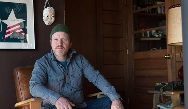 A photo of a man wearing a green cap, sitting in a chair near a mask and a picture of a striped star hanged on the wall.