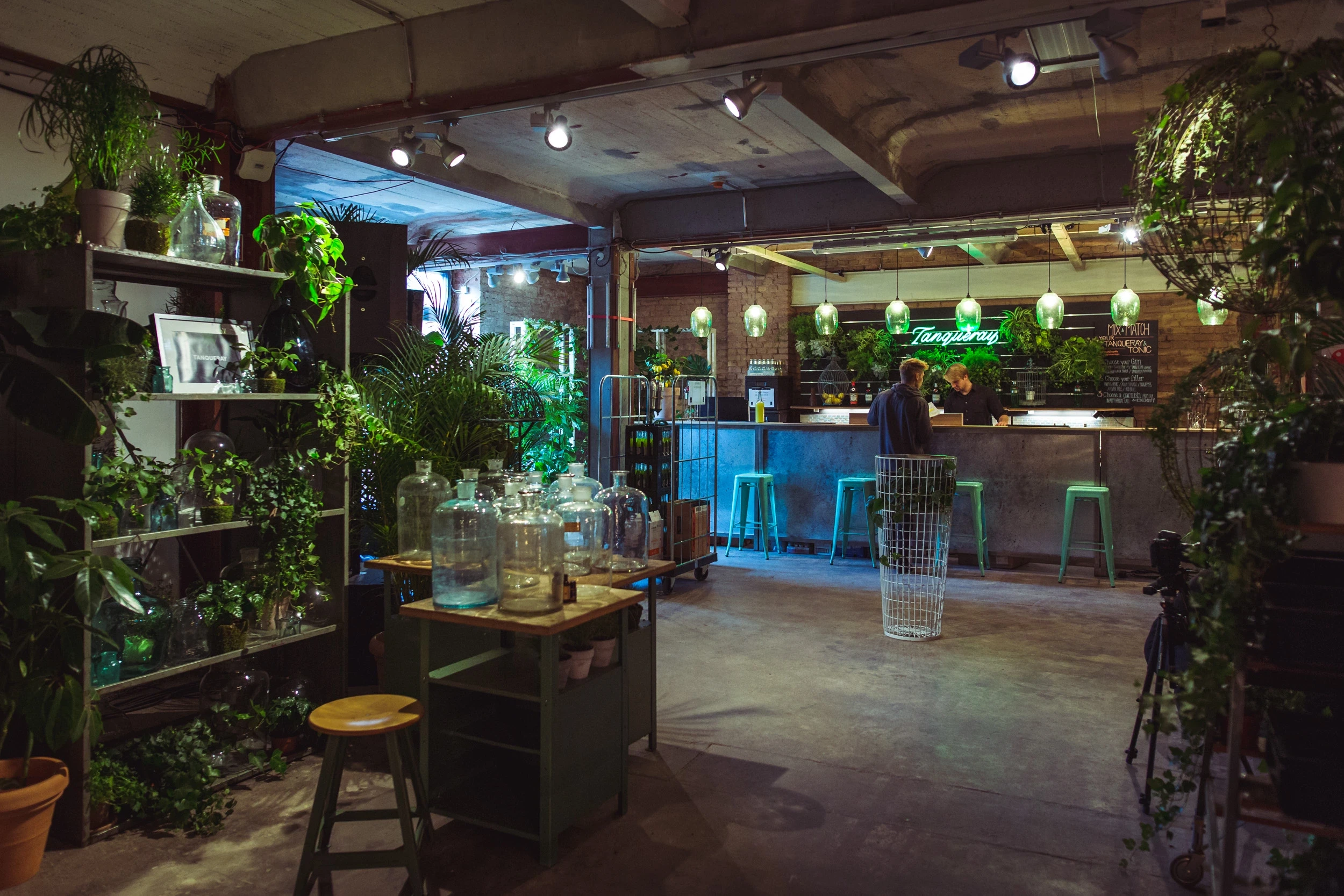 Green Tanqueray bar at DIAGEO BCB18 with neon sign, plant shelves, glass carboys on a workbench and mint-green bar stools.