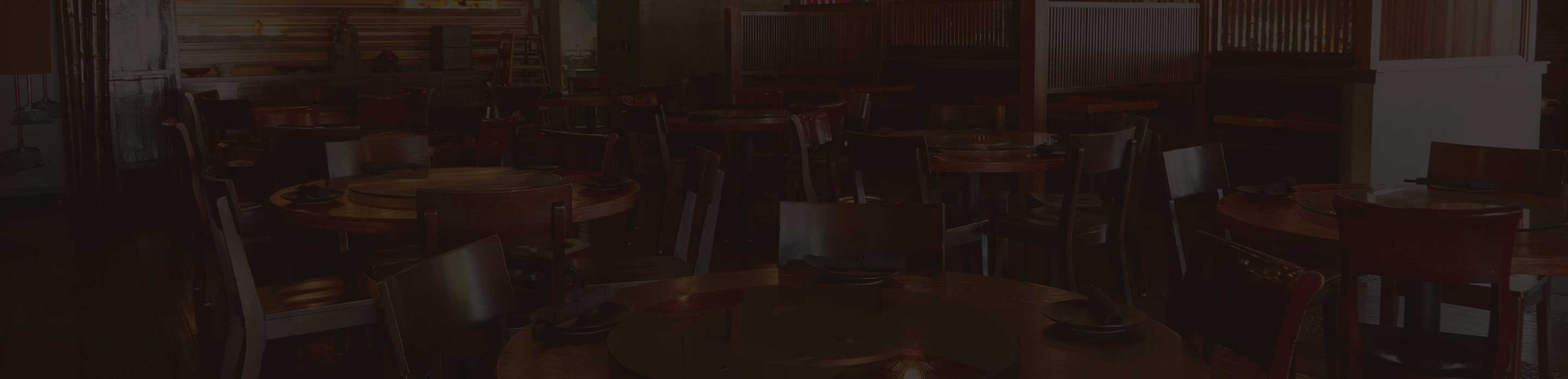 Dimly lit dining area of a restaurant with round wooden tables, dark chairs, and place settings prepared for guests.