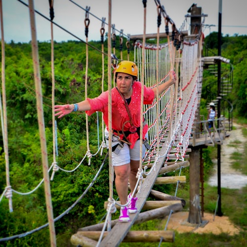 Una persona con casco y arnés cruza un puente de cuerda en un entorno al aire libre exuberante, luciendo concentrada y cautelosa.