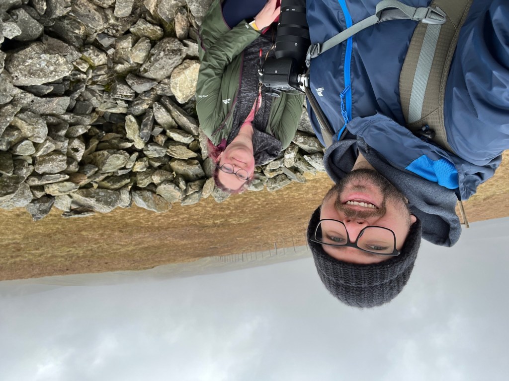 A selfie of Martin and April in the windbreak at Selside Pike. Martin's wearing a blue waterproof and a black wooly hat, whilst April is in her green waterproof.