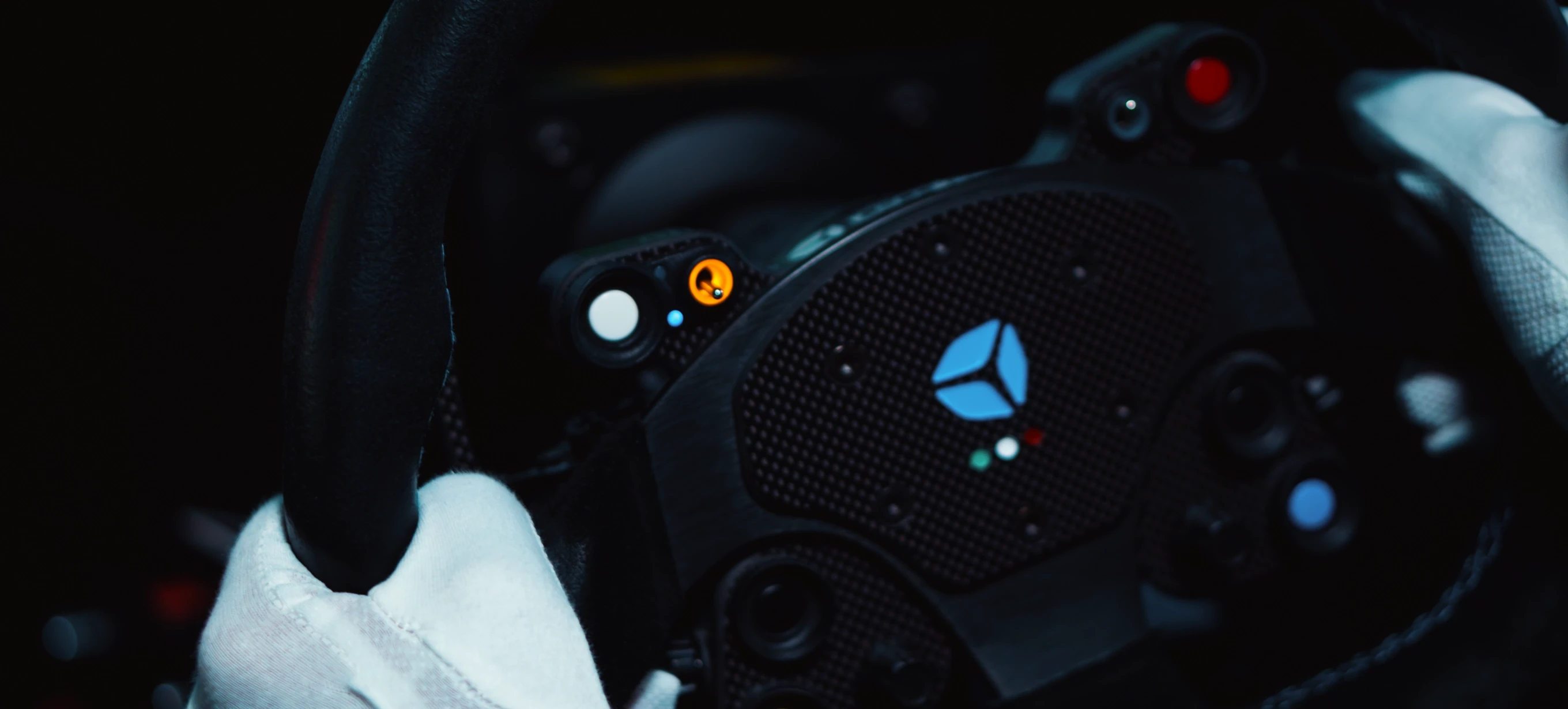 Close-up of a racing simulator steering wheel with illuminated buttons and a blue geometric logo, held by hands in white racing gloves.