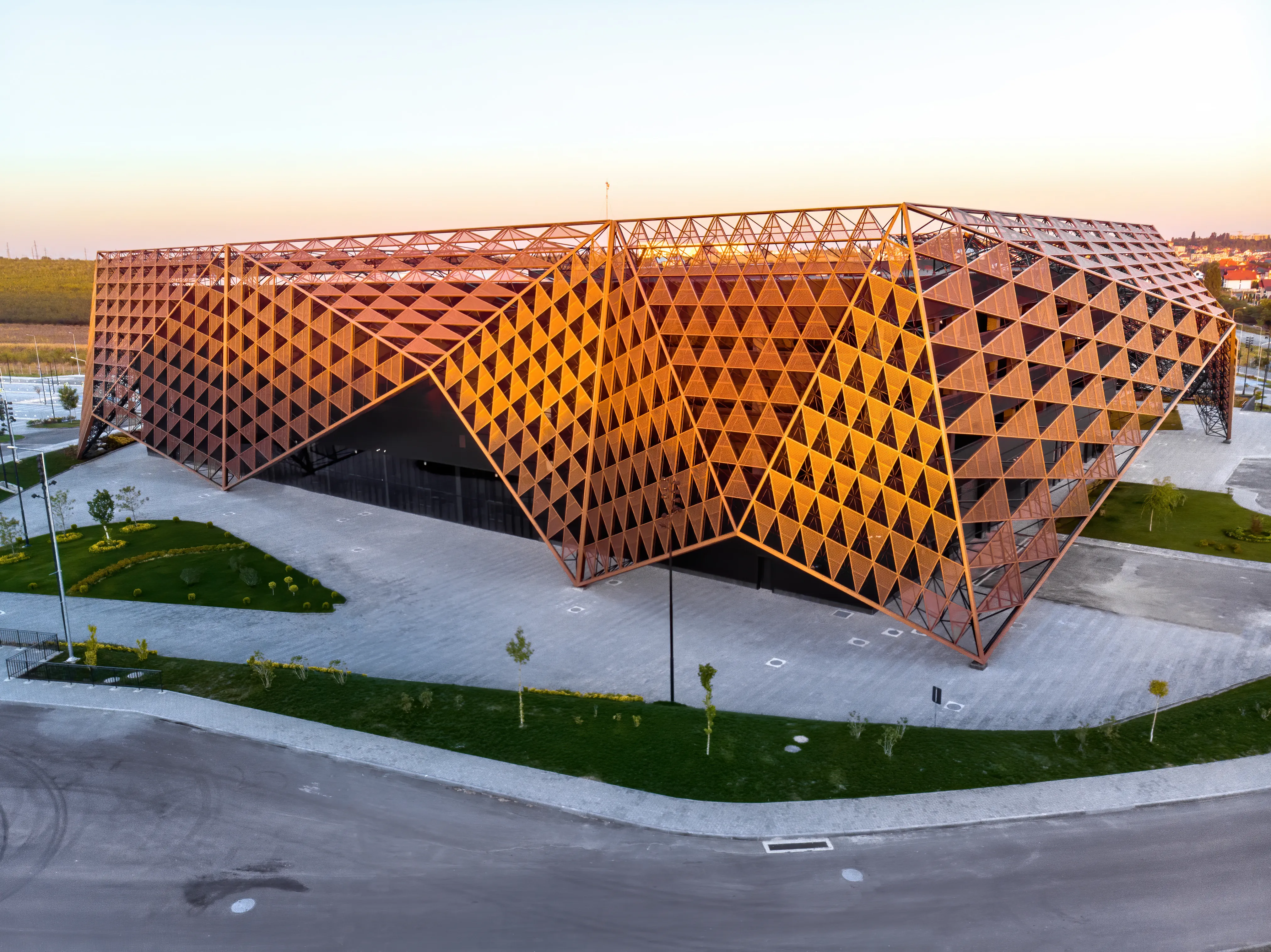 Large-scale architectural pavilion with a complex orange geometric lattice facade and a jagged roofline at sunset.