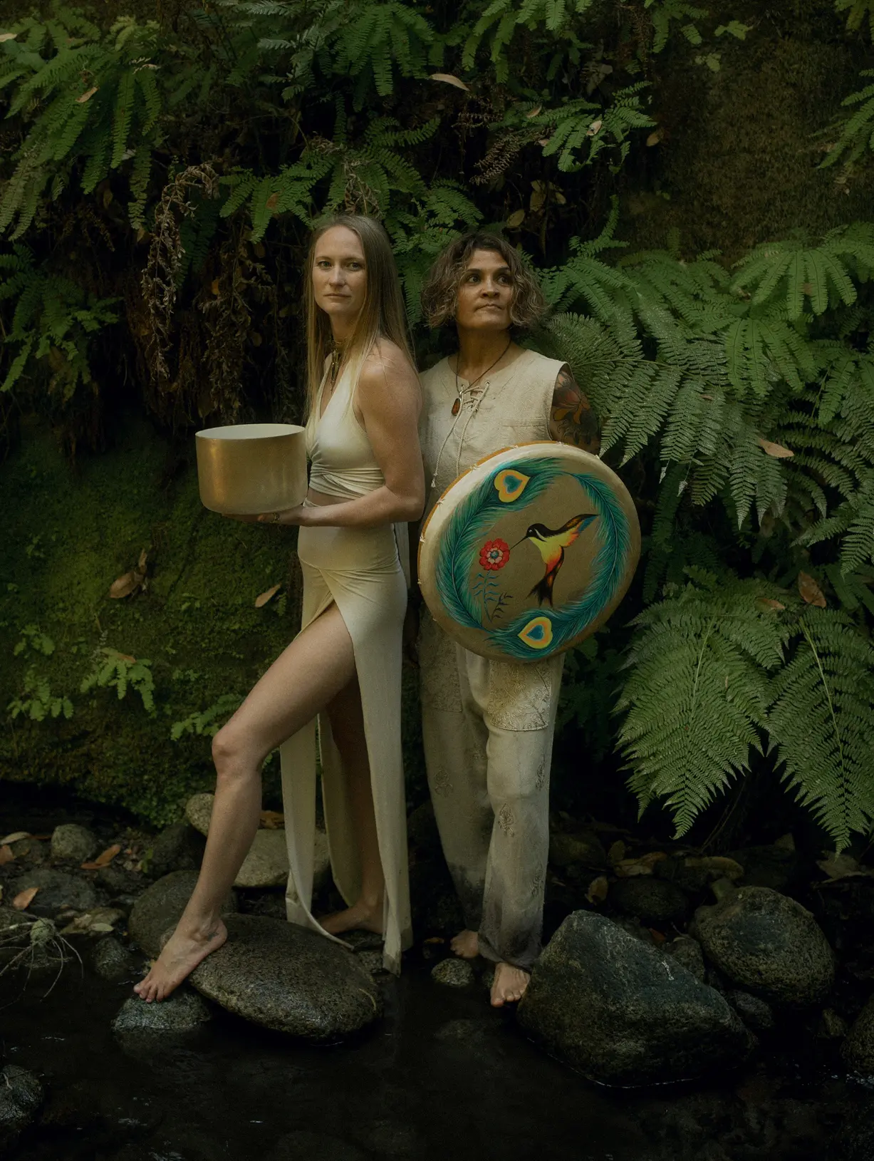 Emeline and Sonia standing back to back in a lush jungle holding a ceremonial drum