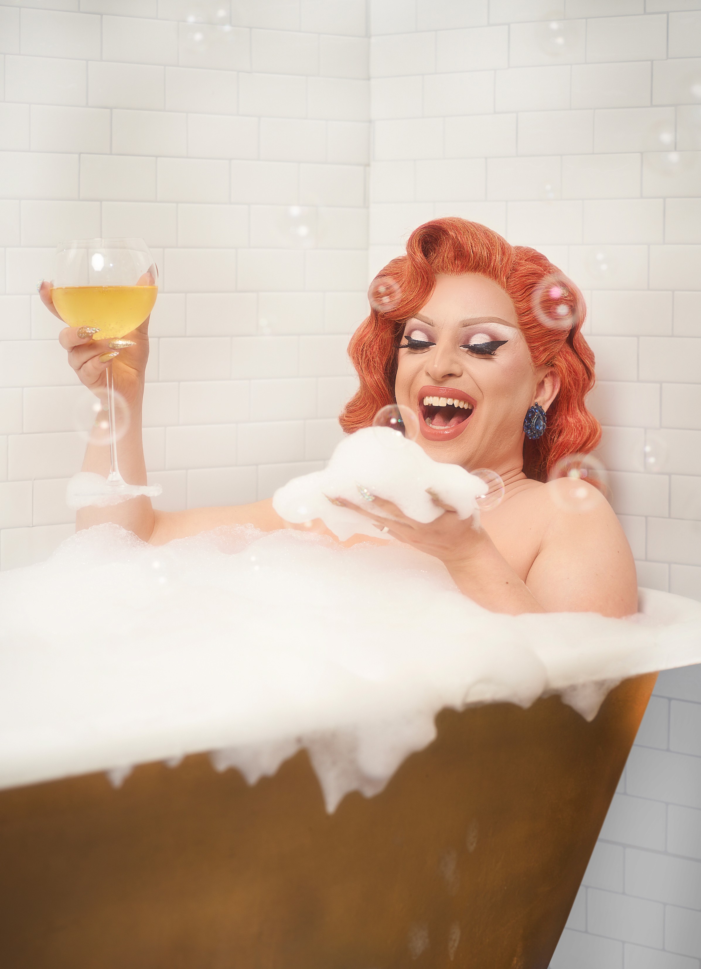 Miz Cracker laughing with a raised wine glass in a foam filled bathtub