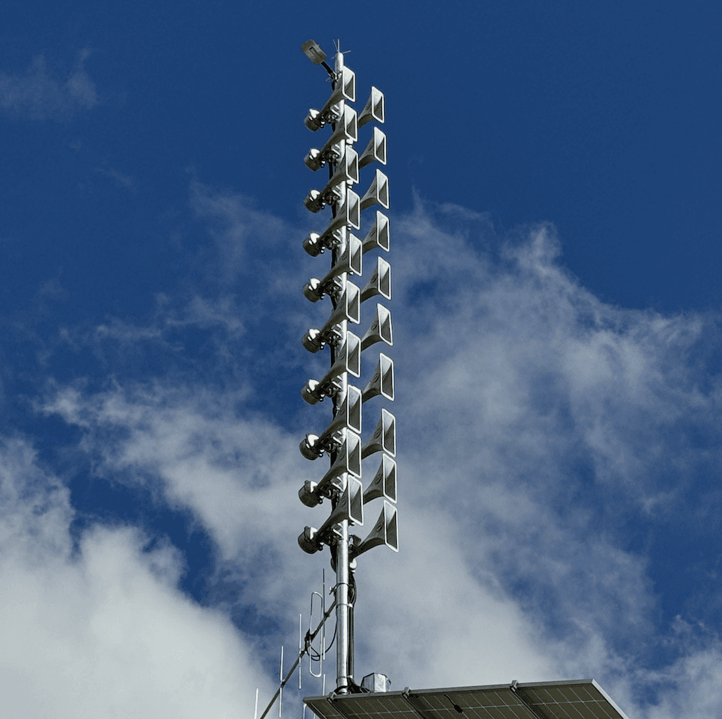 A smiling young man with crossed arms, wearing a plaid shirt and white t-shirt, poses against a dark background.