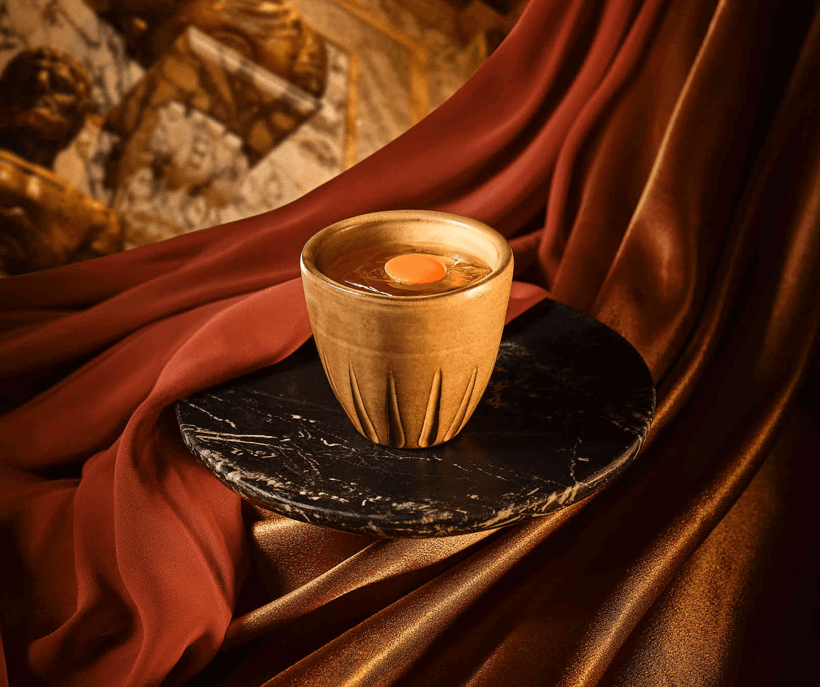 A cocktail in ceramic cup on a dark plate, set against rich, textured fabrics in earthy tones.