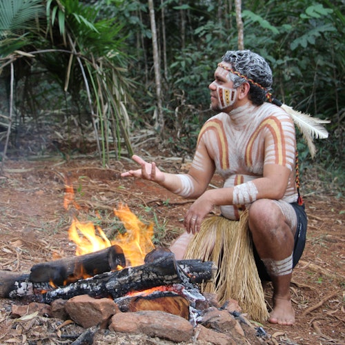 A person with body paint, feathers, and traditional attire sits by a log fire in a forested area, gesturing with their hand.