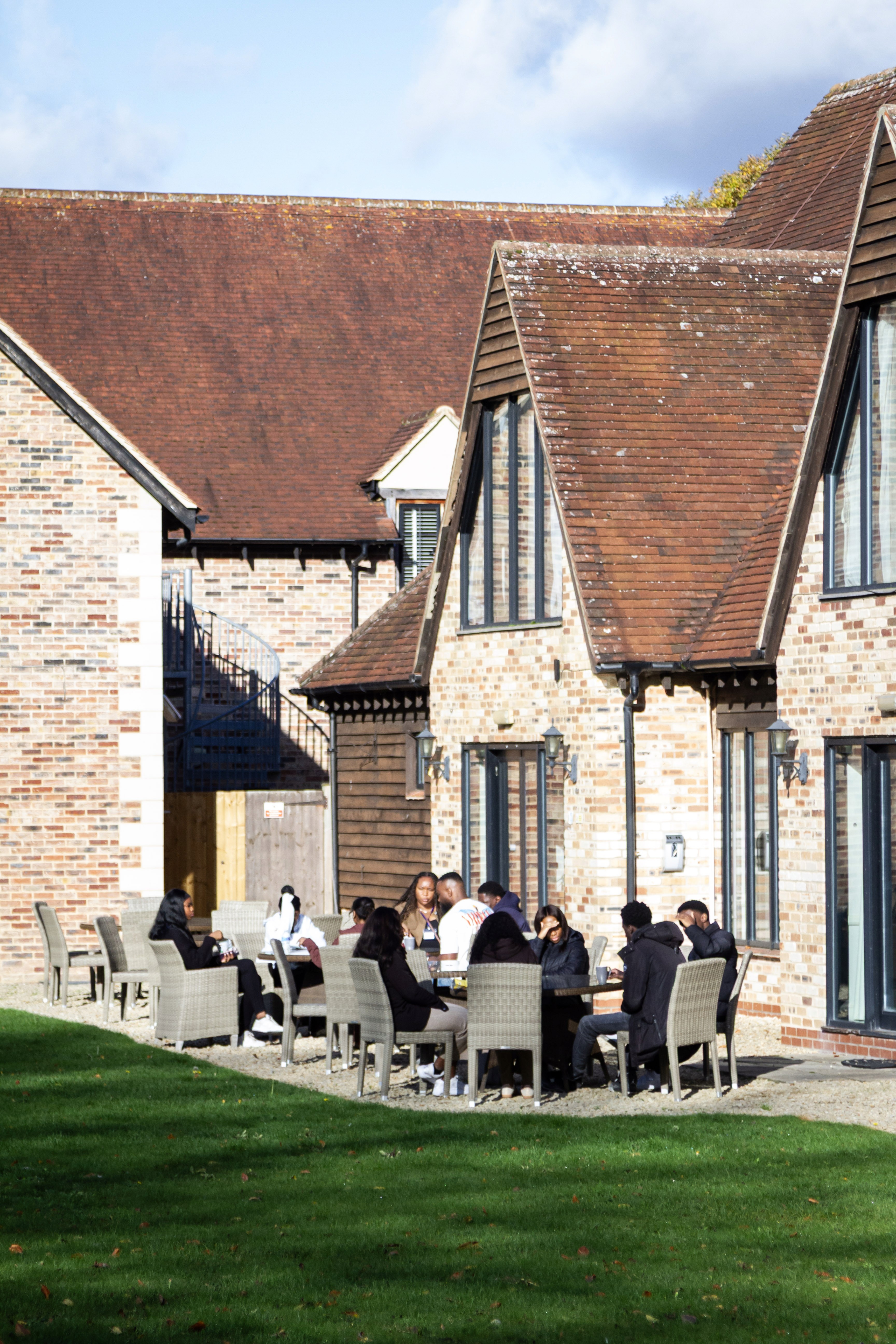 Group of people gathered around tables on a patio with a brick building in the background.