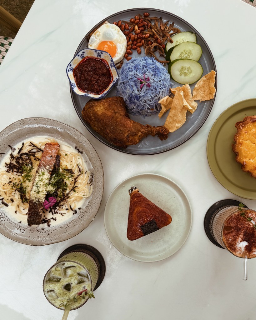 A table setting with two plates of food. One plate features Nasi Kerabu with blue rice, fried chicken, an egg, peanuts, and crackers. The second plate contains pasta with salmon, garnished with herbs. Additionally, there are desserts and drinks, including a green beverage and a layered pastry.