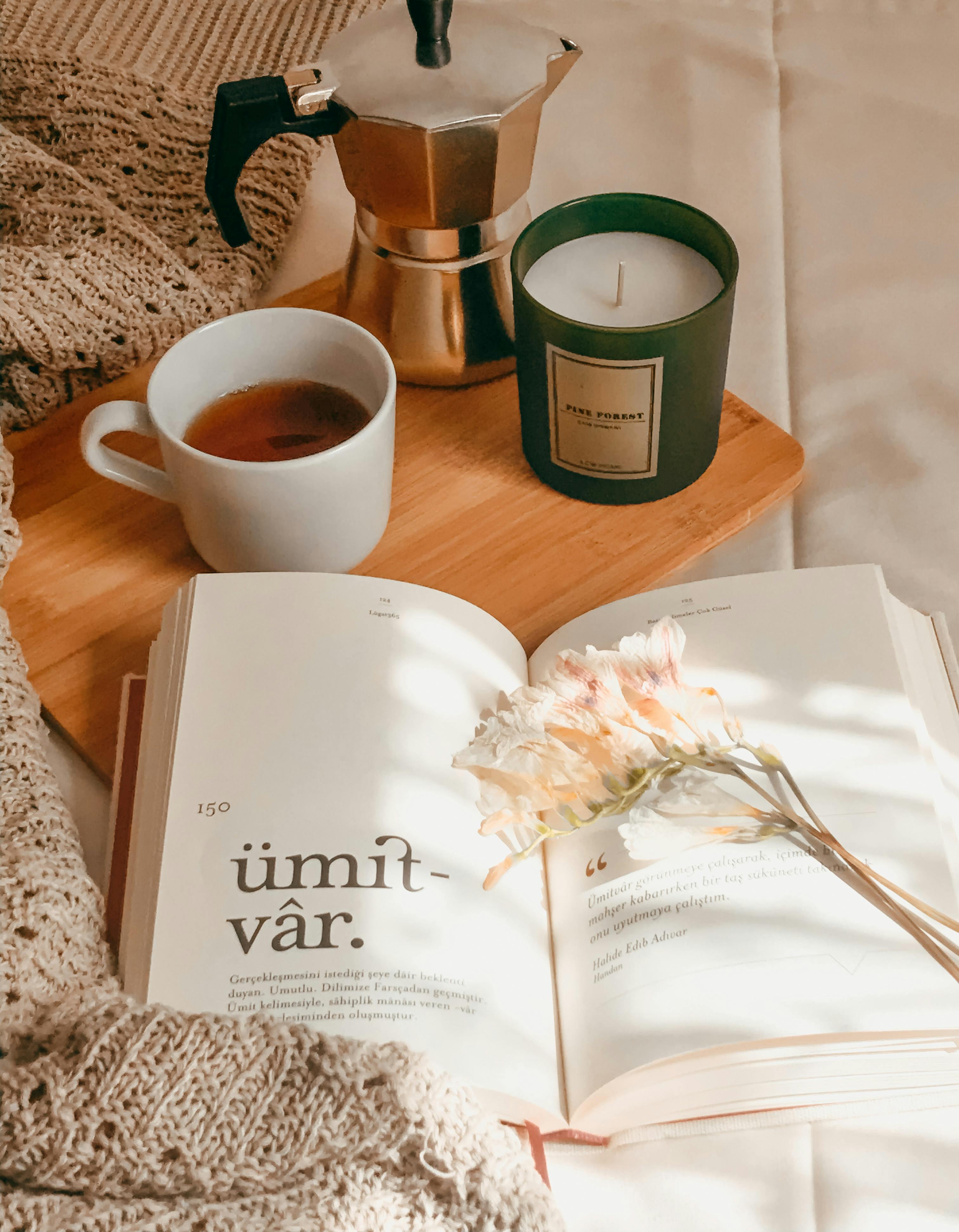 An aesthetic photo featuring an open book, a mug, a candle, and a small teapot placed nearby.