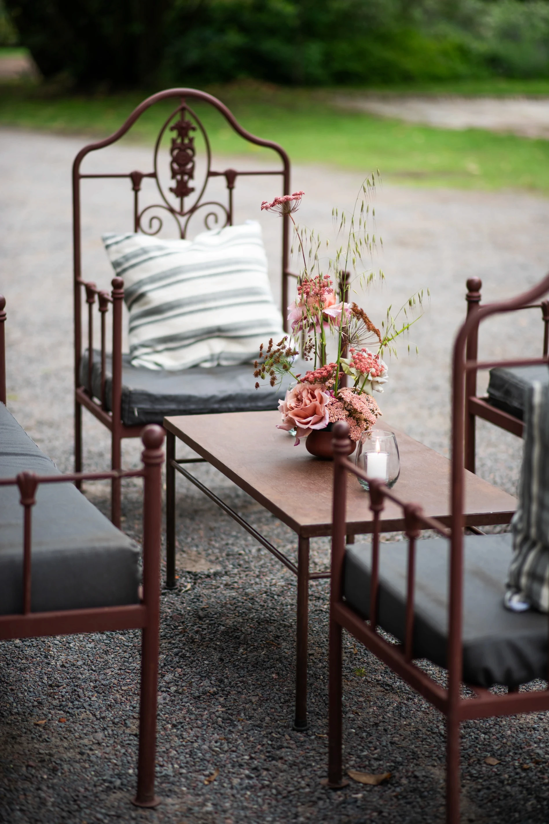 Living de recepción exterior con sillas de hierro forjado, mesa baja de madera y pequeño florero con rosas.