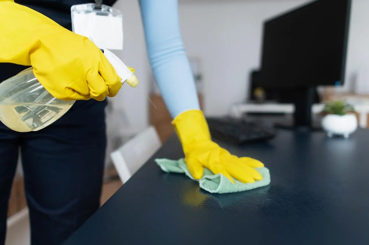 Cleaner wearing yellow gloves wiping desk with spray bottle and cloth