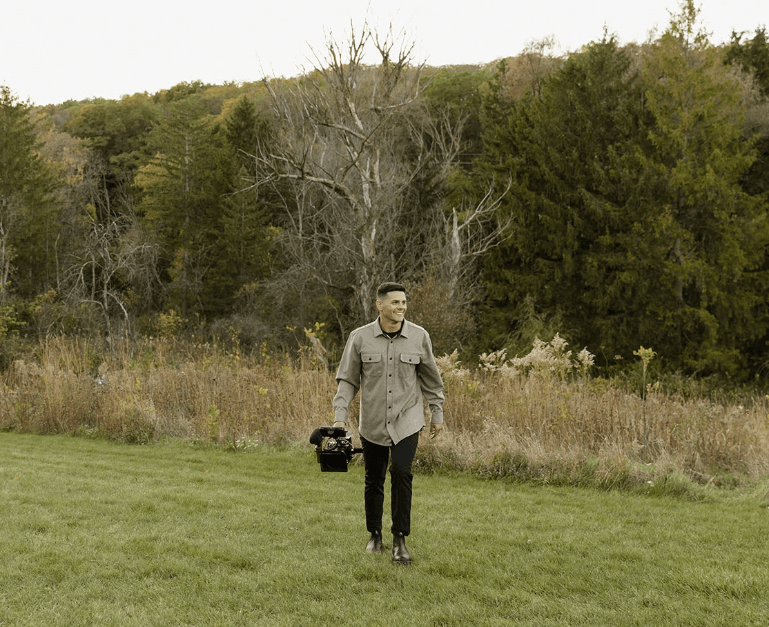 A person carrying a camera walks through a grassy area with trees in the background on an overcast day.