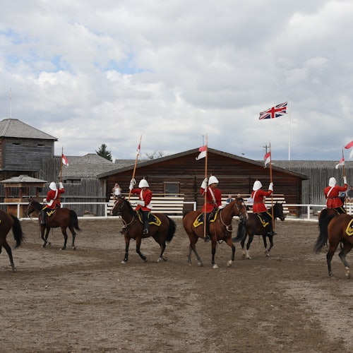 Group of mounted riders in red uniforms parading with flags in a dirt arena, with wooden buildings and British flag in the background.